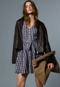 Robe à carreaux bleue, rouge et blanche avec une ceinture à nouer, portée sous une veste en cuir marron foncé. Tenant un sac à main tissé brun texturé.