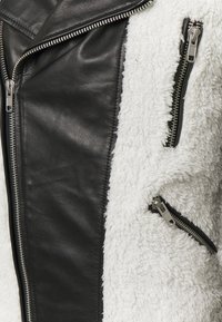 Chaqueta de cuero negro y tela sherpa blanca con cremalleras plateadas en la parte delantera y en los bolsillos, mostrando contraste de texturas y detalles.