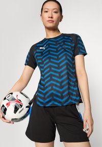 Musthave ja sinine sportlik särk, millel on sakiline muster, valmistatud niiskust eemal hoidvast materjalist, koos mustade lühikeste pükstega. Hoides musterjalgpalli.