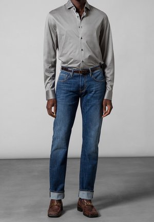 Mężczyzna ubrany w jasnoszarą koszulę na guziki, niebieskie jeansy z podwiniętymi nogawkami i brązowy pasek oraz brązowe skórzane buty na tle szarej podłogi i ściany.