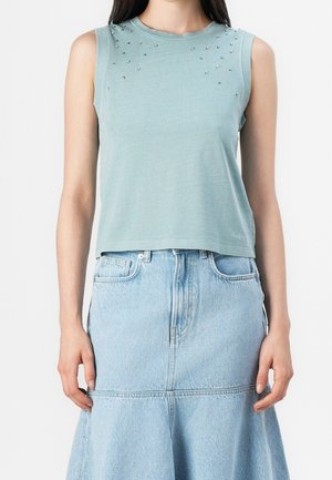Kobieta w bezrękawnikowym, jasnoniebieskim topie z małymi srebrnymi ćwiekami przy dekolcie, w połączeniu z jasną, długą do talii spódnicą z denimu.