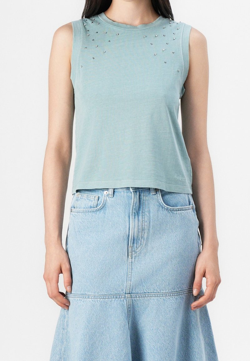 Femme portant un haut sans manches bleu clair avec de petits clous argentés près de l'encolure, associé à une jupe en denim clair taille haute.