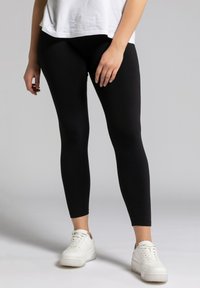 Leggings noirs en matériau extensible, design ajusté, associés à des baskets blanches, texture lisse et sans motifs ni accents notables.