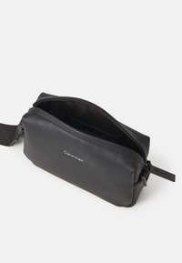 Pochette en cuir noir texturé avec fermeture à glissière, arborant un logo sur le devant et une forme rectangulaire compacte. L'intérieur est doublé.