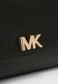 Musta nahkalaukku, jossa on silmiinpistävä kulta "MK" -logo korostuksena, esittelee sileän pinnan ja siistit linjat.