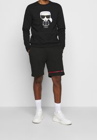Felpa nera con una figura cartoon che indossa occhiali da sole e un completo, abbinata a pantaloni corti neri con una striscia rossa e sneakers bianche.