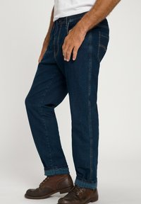 Jeans de mezclilla azul con corte recto, que presentan un patrón de cuadros en el dobladillo y zapatos de cuero marrón. Costuras visibles en los laterales.