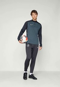 Nike sportinė apranga, kurios aprašyme yra tamsiai pilka, ilgomis rankovėmis, su užtrauktuku ir musta priglundančios kelnes su logotipu. Laikoma futbolo kamuolys, juodi batai.