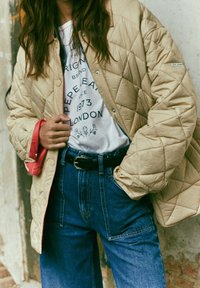 Chaqueta acolchada beige con cierre de botones a presión sobre una camiseta blanca con estampado gráfico, combinada con unos vaqueros de mezclilla azul oscuro de talle alto y un cinturón negro.