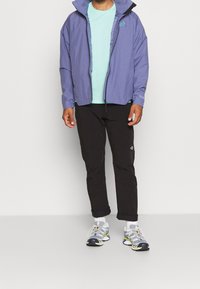 Fialová nepromokavá bunda s nastavitelnou kapucí, doplněná světle modrým tričkem, černými kalhotami a šedými sportovními botami s barevnými akcenty.