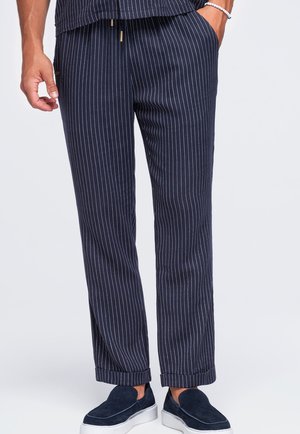 Homme portant un pantalon bleu marine à fines rayures, retroussé aux chevilles, avec des chaussures bleu marine sans lacets et un bracelet de perles au poignet.