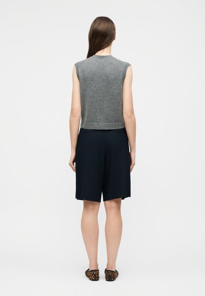 Γυναίκα με μακριά καστανά μαλλιά, φορώντας γκρι αμάνικο τοπ, μαύρα σορτς μέχρι το γόνατο και παπούτσια με λεοπάρ σχέδιο, στάσιμη με πλάτη προς τα εμπρός.