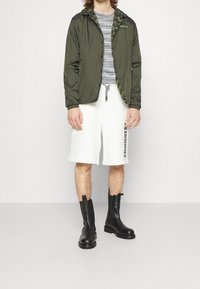 Chaqueta ligera de color verde oliva con un forro de patrón zigzag. Combinada con pantalones cortos blancos y botas negras hasta el tobillo. Se puede ver una blusa de rayas horizontales.