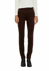 Pantalon slim en velours côtelé marron à texture lisse, doté de cinq poches et d'une braguette zippée, porté avec un haut blanc à manches longues et des bottes noires.