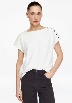 Frau mit zurückgebundenem Haar, trägt ein weißes kurzärmeliges Oberteil mit schwarzen Knöpfen an der Schulter und schwarze Hosen, Hand in der Tasche.