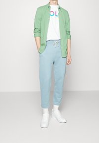 Zielona koszula z guzikami, biała koszulka z grafiką, jasnoniebieskie dresy i białe sneakersy. Casualowy strój z miękkich tkanin i luźnym krojem.