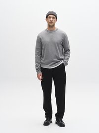 Grijs shirt met lange mouwen en ronde hals, zwarte broek en een grijze gebreide muts. Man staat rustig met handen in broekzakken.