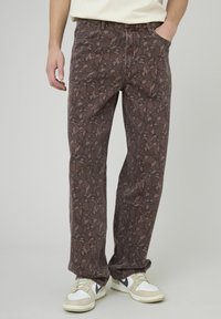 Jean marron à motifs avec une coupe ample, doté d'un tissu texturé, d'une ceinture et de deux poches latérales. Porté avec des baskets blanches et beige.