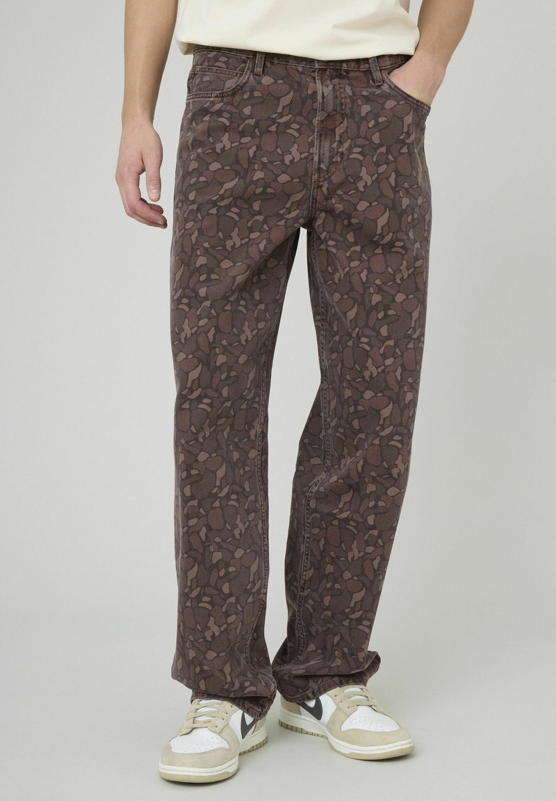 Jean marron à motifs avec une coupe ample, doté d'un tissu texturé, d'une ceinture et de deux poches latérales. Porté avec des baskets blanches et beige.