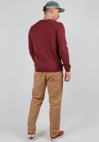 Pull à manches longues bordeaux avec un col rond, associé à un pantalon chino beige et des baskets brunes, sur un fond clair uni.