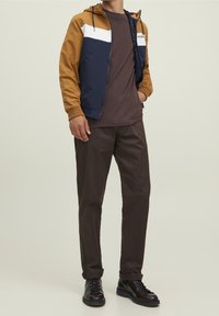 Jacket v barevném bloku v hnědé, námořnické a bílé; hnědé tričko pod ním; tmavé kalhoty; černé šněrovací boty; ležérní design.