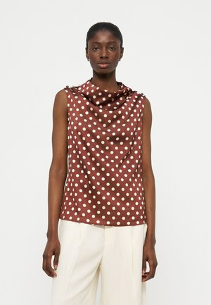 Γυναίκα που φοράει αμάνικο καφέ τοπ με λευκά πουά και ντραπαρισμένο ντεκολτέ, συνδυασμένο με άνετο λευκό παντελόνι, στέκεται μπροστά σε απλό φόντο.