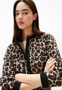Cardigan med leopardmønster og svart kant, strikket tekstur og knappefester. Har ribbestrikkede mansjetter og en subtil merkeaccent på ermet.