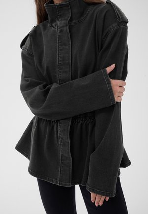 Personne portant une veste en jean noire oversized avec de larges manches et un col haut, croisant un bras sur son corps.