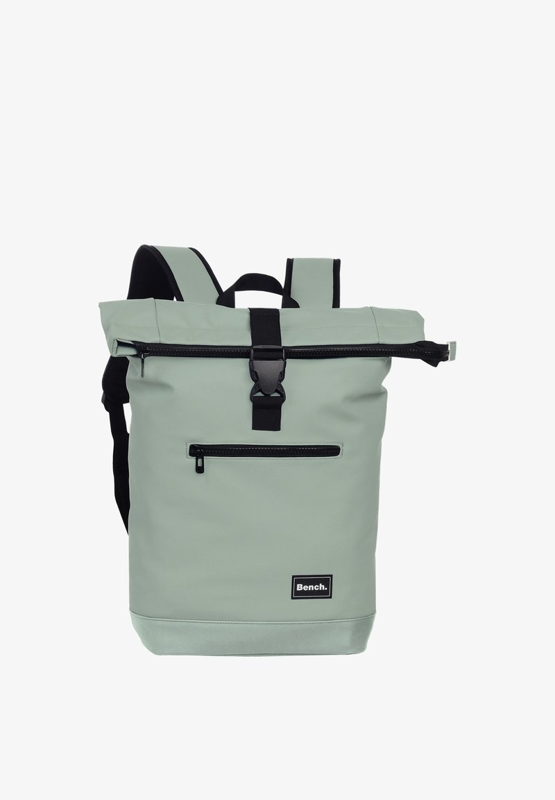 Sac à dos à rabat enroulable gris clair avec poches zippées noires, fermeture à boucle, bretelles rembourrées, et un patch logo "Bench." sur le devant.