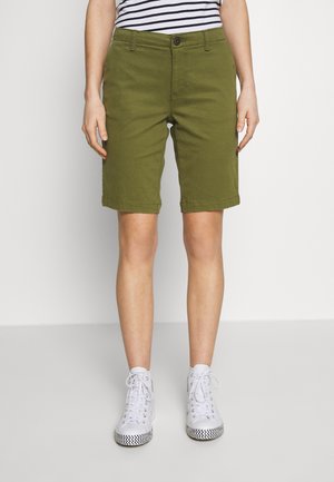 Personne portant un short olive mi-genou, des baskets montantes blanches avec des semelles à motifs, et un t-shirt à rayures noir et blanc.