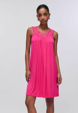 Kobieta w jaskrawo różowej sukience bez rękawów z okrągłym dekoltem ozdobionym ćwiekami, stojąca na tle jasnego, jednolitego tła.