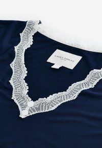 Îmbrăcăminte din bumbac de culoare bleumarin, cu dantelă albă la gulerul în formă de V, prevăzută cu două butoane decorative și o etichetă a brandului pe spate.