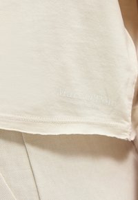 Κοντινό πλάνο μπεζ υφάσματος με λεπτό λογότυπο "Marc O' Polo" ραμμένο κοντά στο τελείωμα, που φοριέται πάνω από ανοιχτόχρωμο παντελόνι με υφή.