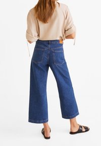 Jeans de denim azul de pierna ancha con una cintura alta, que presentan bolsillos de parche y costuras en contraste. Combinados con una blusa beige y sandalias negras.