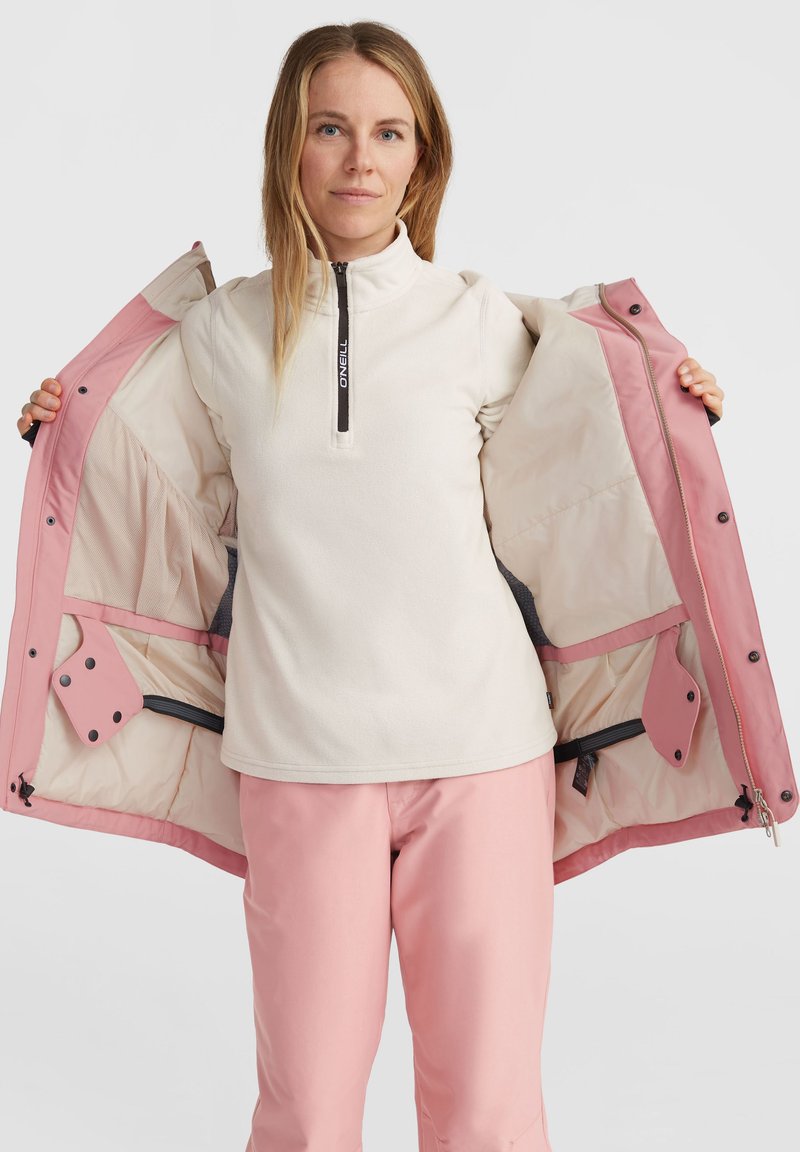 Vrouwenjack met een roze buitenlaag, een crèmekleurige fleece binnenlaag, een kwartrits hals en meerdere binnenzakken. Zachte, gladde textuur.