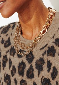 Collier chaîne ton or superposé avec une chaîne à maillons supplémentaires, porté sur un pull beige à motifs avec des taches léopard noires.