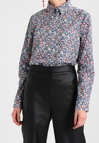 Femme portant une chemise à boutons florale ajustée rentrée dans un pantalon en cuir noir taille haute, se tenant devant un fond uni.