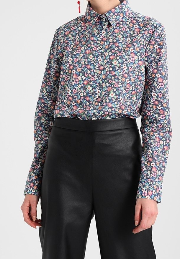 Femme portant une chemise à boutons florale ajustée rentrée dans un pantalon en cuir noir taille haute, se tenant devant un fond uni.