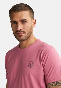 Rosa Baumwoll-T-Shirt mit rundem Halsausschnitt und einem kontrastierenden schwarzen Logo-Print auf der linken Brust. Kurze Ärmel und normale Passform.