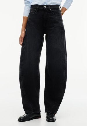 Person iført højtaljede vidde sorte jeans, lyseblå sweater, sorte læder loafers og et gyldent armbånd med den ene hånd i lommen.