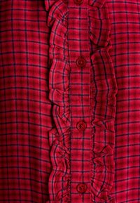 Red plaid shirt with a ruffled front, featuring red buttons and a textured fabric. The plaid pattern includes blue lines against the red background.