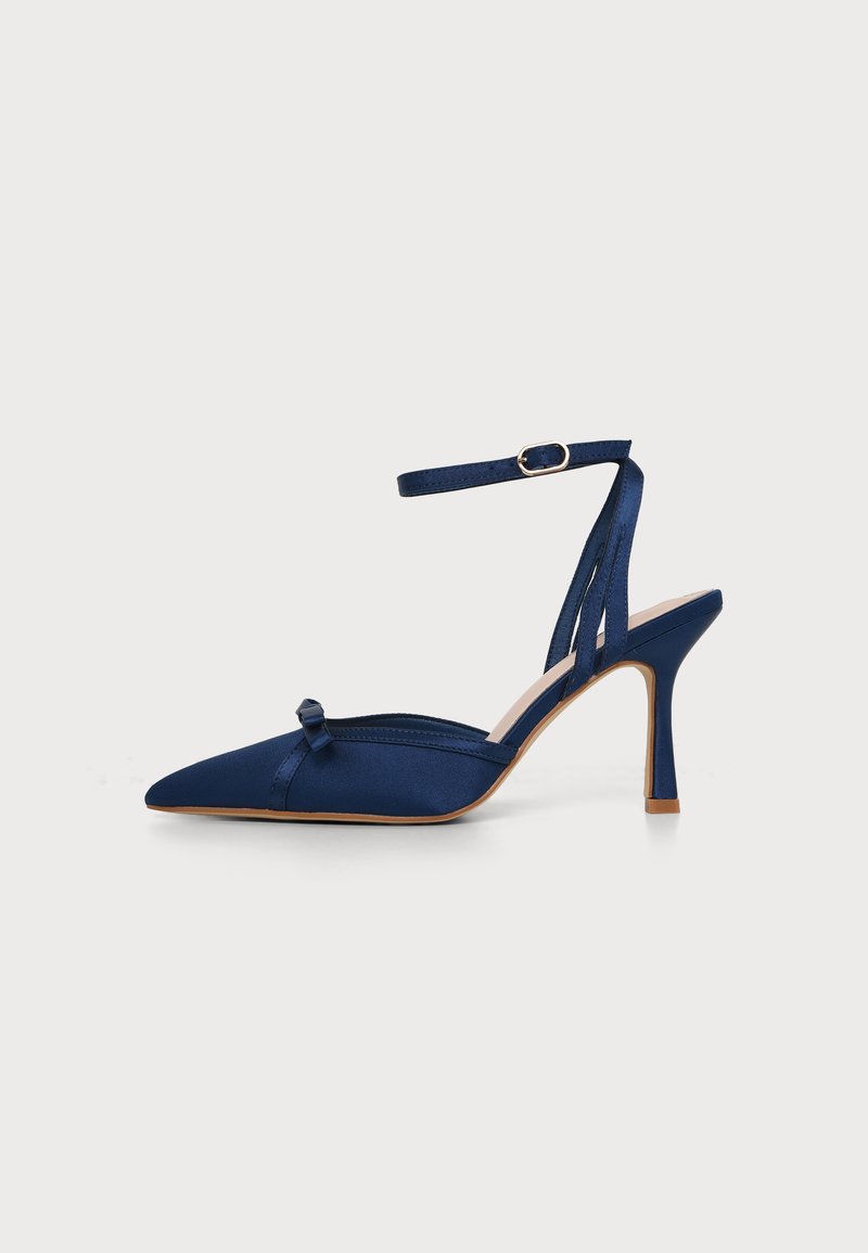 Chaussure à talon haut en satin bleu marine, bout pointu, bride de cheville avec boucle et petit nœud décoratif à l’avant.