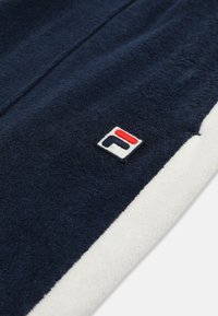 Ręcznik w granatowo-białych kolorach z małym haftowanym logo w kolorach czerwonym, białym i granatowym przy brzegu na jasnym tle.