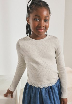 Ung pige med flettet hår iført en lys beige langærmet top og en blå nederdel, stående indendørs mod en ensfarvet baggrund.