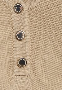 Beige strikket stof med tre sorte, blanke knapper med præget tekst. Teksturen er ribbet og fremviser et subtilt mønster.