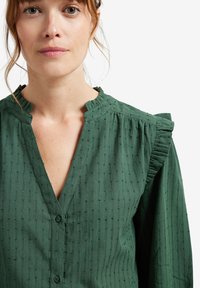 Green button-up shirt with a textured fabric, featuring a V-neck, ruffled collar, and subtle vertical stripes.