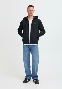 Svart hettejakke med glidelås og snor, to frontlommer, båret over en hvit T-skjorte, kombinert med lyseblå jeans og svarte sneakers.