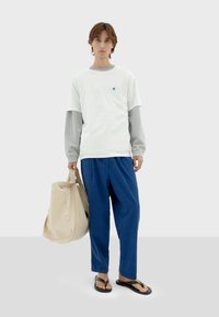 Casual outfit met een wit en grijs langslaved shirt, blauwe gestructureerde broek en zwarte slippers, met een beige boodschappentas.