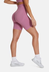 Kobieta w fioletowych, wysokich szortach rowerowych i białych sportowych sneakersach stoi z jedną nogą lekko uniesioną na jednolitym tle.