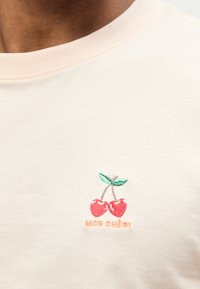 Πουκάμισο εκρού με κεντητό σχεδιασμό δύο κόκκινων κερασιών και πράσινα φύλλα πάνω από πορτοκαλί κείμενο που γράφει "MON CHÉRI" στο στήθος.
